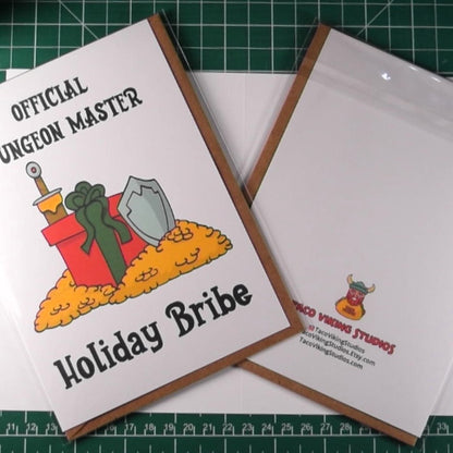 The 'Official Dungeon Master Holiday Bribe' greeting card displayed on a crafting table, surrounded by various colored dice, giving a close-up of both the front design and the transparent protective sleeve packaging.